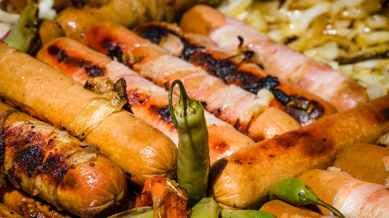 A variety of grilled hot dogs on a street cart with bacon, onions, and peppers