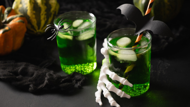Two green Halloween drinks, one gripped by skeleton hand and with a bat straw and the other with a fake spider on edge of glass