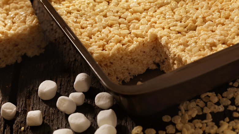 Corner of Rice Krispies treat cake inside a pan, with marshmallows on the dark, wooden table