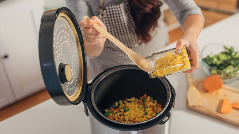 Woman's hands pouring corn and other ingredients into a slow cooker while preparing food in the kitchen.