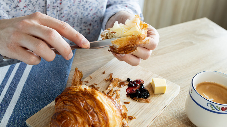 Hands buttering ripped up croissant next to cup of coffee