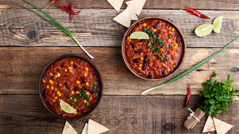 two bowls of chili on a wooden table with produce