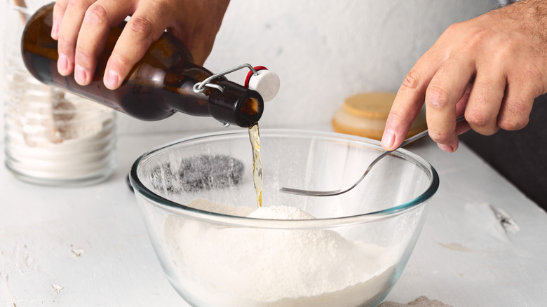 A person adding beer from a bottle to a glass bowl with flour in it.
