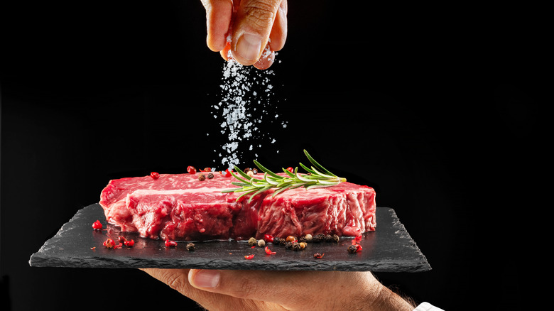 A hand salting a raw ribeye steak on a black plate.