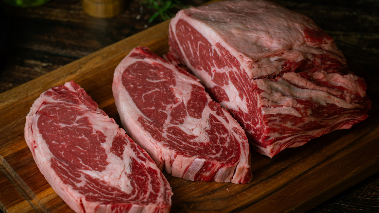 Two slabs of raw ribeye cut off of a large chuck of meat on a wooden cutting board