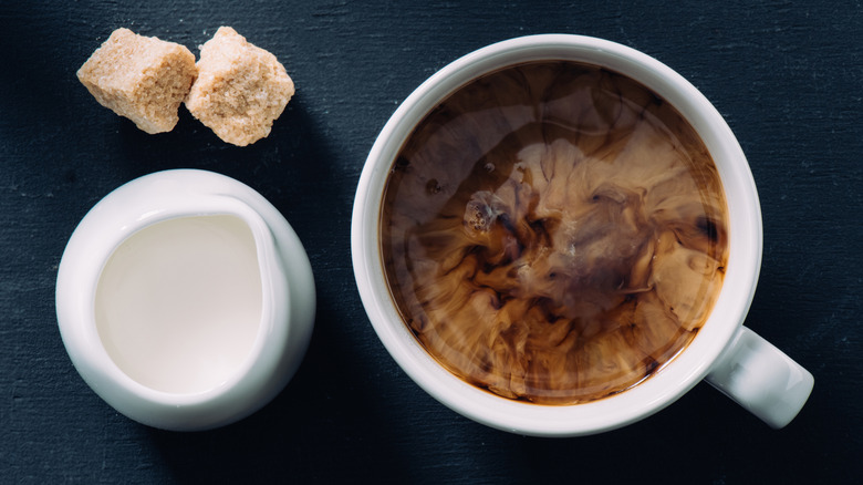 Two brown sugar cubes in front of cream next to white mug with cream spreading through as an overhead shot