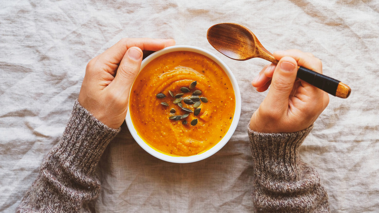A bowl of tomato soup with pumpkin seeds, being held by a pair of hands holding a wooden spoon