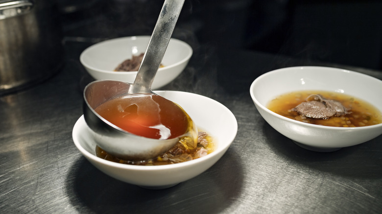 Consommé being ladled into a white bowl with meat slices inside, with another a filled bowl next to it on a metal surface