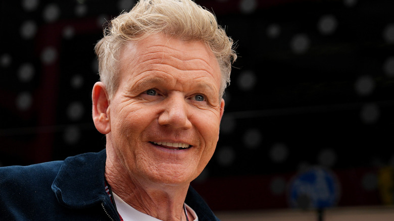 Close-up of Gordon Ramsay wearing a white chef's coat in front of a promotional background