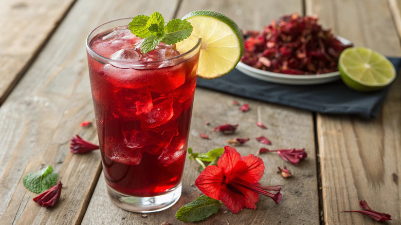 Glass of red iced tea with mint and lime next to hibiscus flower and loose tea leaves