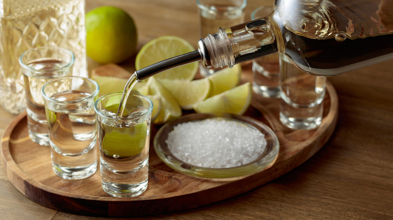 Tequila being poured into a shot glass on a tray with other shots, salt, and lime wedges