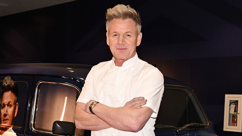 Gordon Ramsay with his arms folded standing in front of a car with a Gordon Ramsay decal on it.