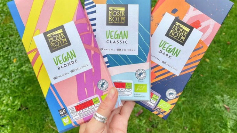 Holding three Moser Roth Vegan Chocolate Bars in colorful wrappers against a green grass background