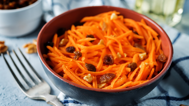 Shredded carrots and raisin salad with small walnut pieces in a brown bowl with the top of a fork next to it on a blue and white kitchen towel