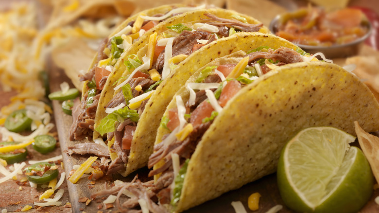 Three hard shell tacos with shredded beef, cheese, lettuce, and tomato near a cut lime on a wooden surface