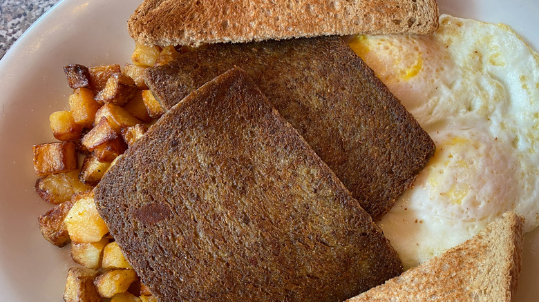 A classic diner breakfast plate featuring two browned slices of fried scrapple, crispy diced potatoes, over-easy eggs, and buttered wheat toast.