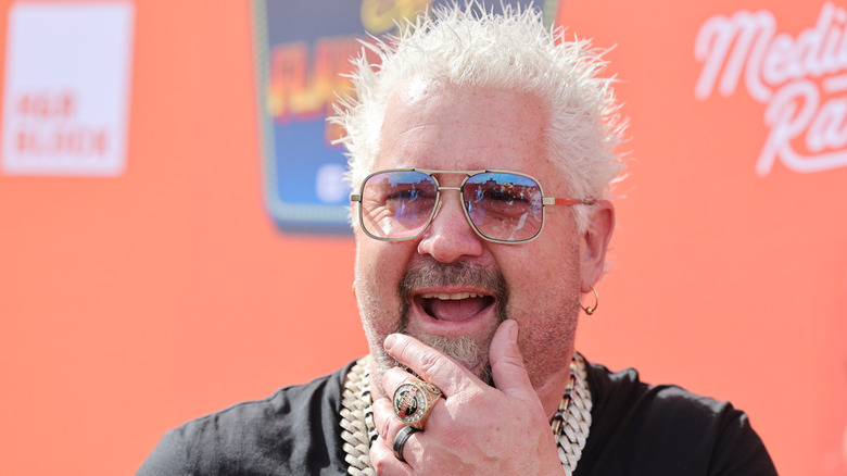 Guy Fieri smiling with his hand on his chin, wearing sunglasses, gold jewelry, and a black shirt in front of an orange 