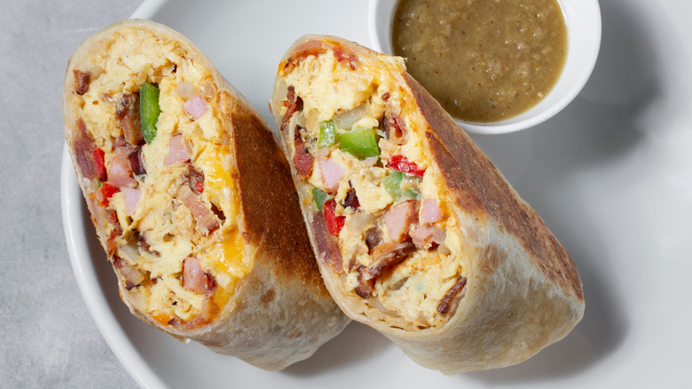 Breakfast burrito cut in half on white plate with a pot of sauce