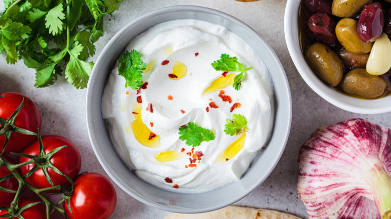 Whipped feta cheese dip in a gray bowl surrounded by olives, garlic, tomatoes, and herbs