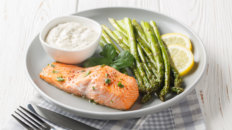 A plate of cooked salmon, asparagus, and lemon with a bowl of tartar or mayo-based sauce