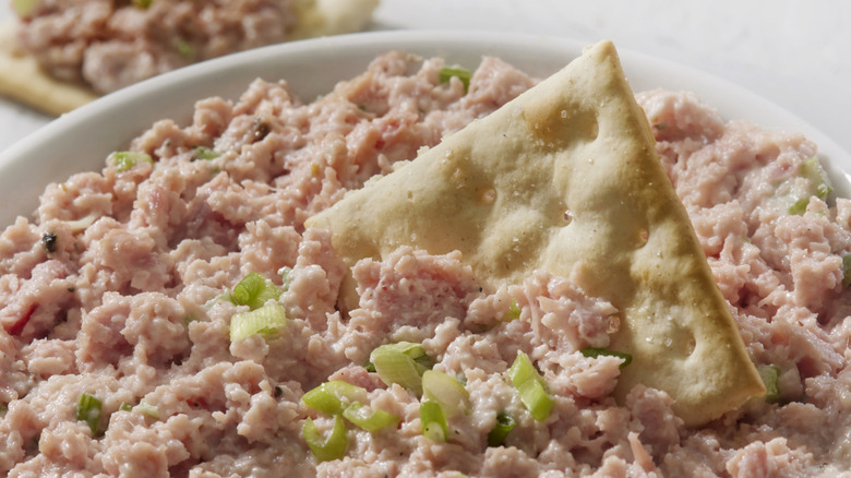 A bowl of bologna salad topped with scallions with a saltine cracker