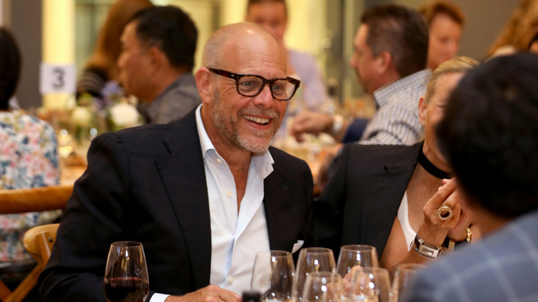 Alton Brown sitting down at a large gathering with glasses of wine in front of them.