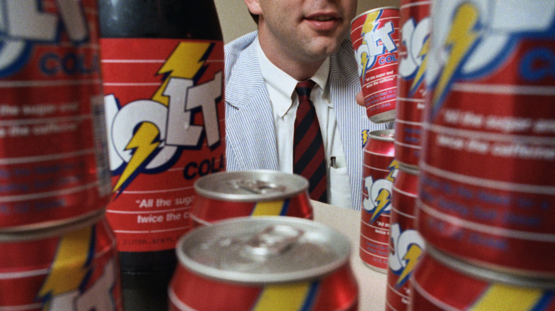 A man in a suit and tie poses surrounded by bottles and cans of Jolt Cola.