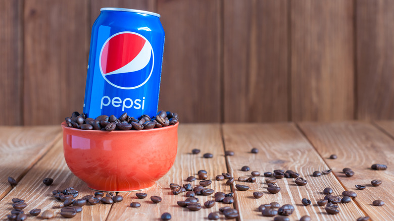 A can of Pepsi placed inside a red bowl filled with coffee beans, with more beans scattered across a wooden table.