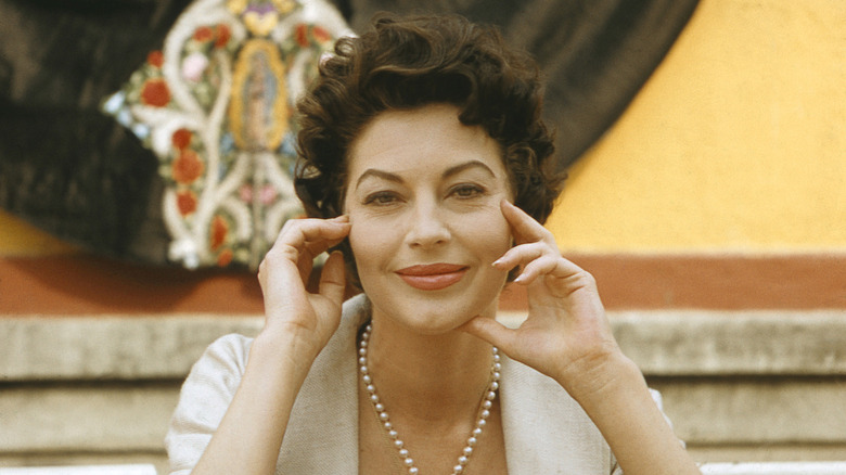 Ava Gardner smiles for photo wearing pearl necklace and hands on face