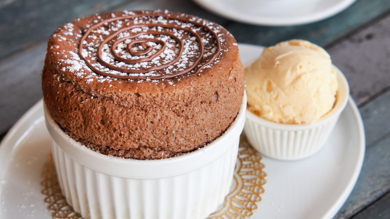 A chocolate soufflé sitting in a white ramekin on a white plate with butter or maybe ice cream nearby.
