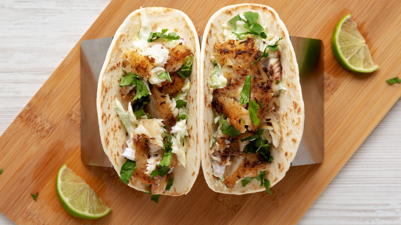 Two tacos on a platter filled with fish, herbs, and fresh lime