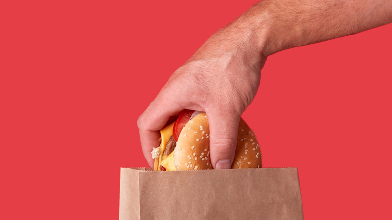 Hand pulling a burger out of a brown paper bag against a salmon pink background.
