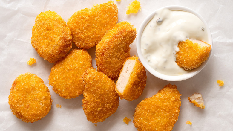 Several chicken nuggets scattered on a white surface, with one cut half in a white sauce in a small white bowl