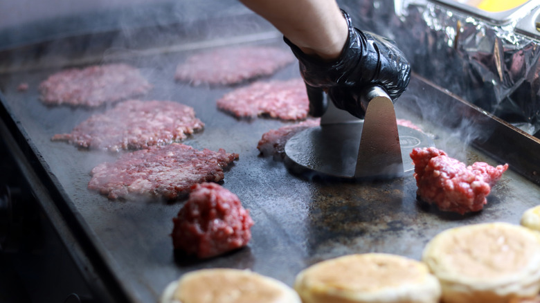 Smashburgers on a grill with a gloved hand pushing a press down on one of them