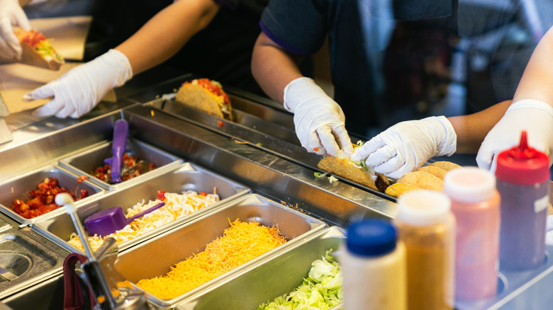 Three white-gloved workers make tacos from ingredient pans