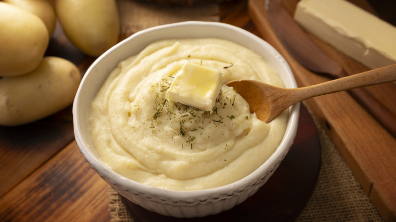A bowl of mashed potatoes with butter and a wooden spoon with ingredients in the background.