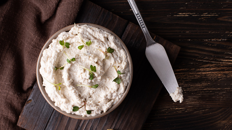 Cream cheese with chives in a bowl on a table with a knife nearby.
