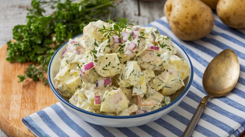 Bowl of potato salad with herbs and red onion on cutting board with spoon.