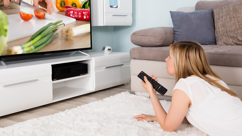 A woman lays on the floor watching a cooking show on TV