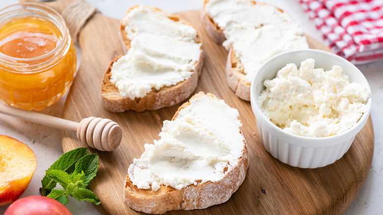 Ricotta spread on three pieces of toasted Italian bread, with a small ceramic white bowl of ricotta on a wooden board, and orange jam on the side