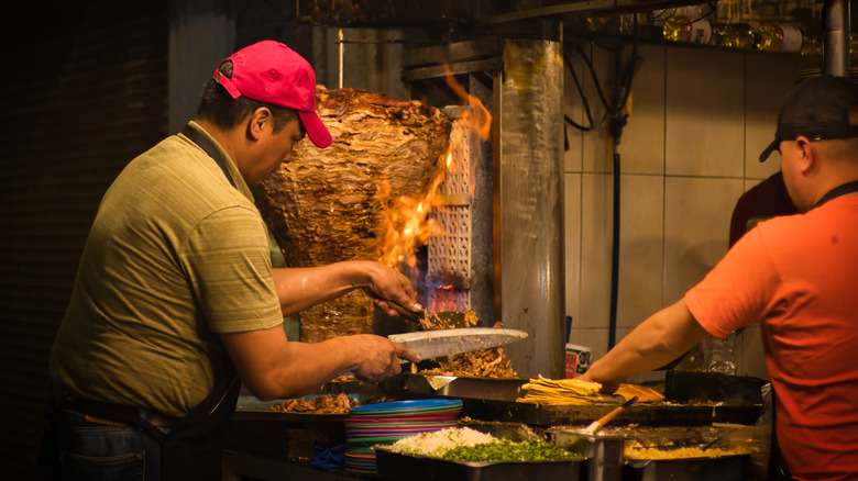 A cook uses a butcher knife to cut slices from pork on a spit