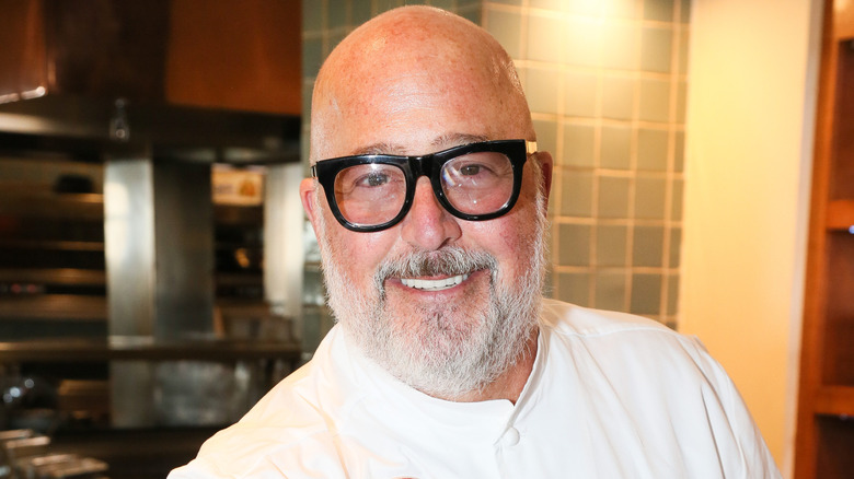 Andrew Zimmern smiling and wearing a white chef jacket in a kitchen