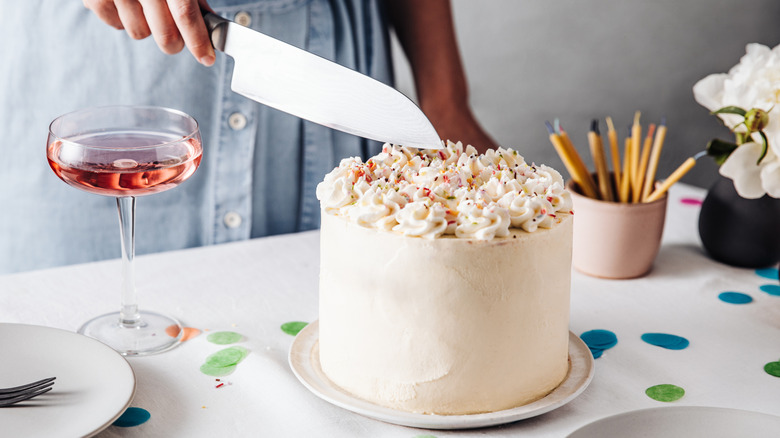 Frosted white birthday cake with with rainbow sprinkles on top and person with chef's knife about to slice through. Cocktail glass with pink drink inside to the left