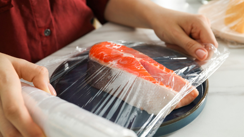 A person stretches plastic wrap over fresh salmon on a plate