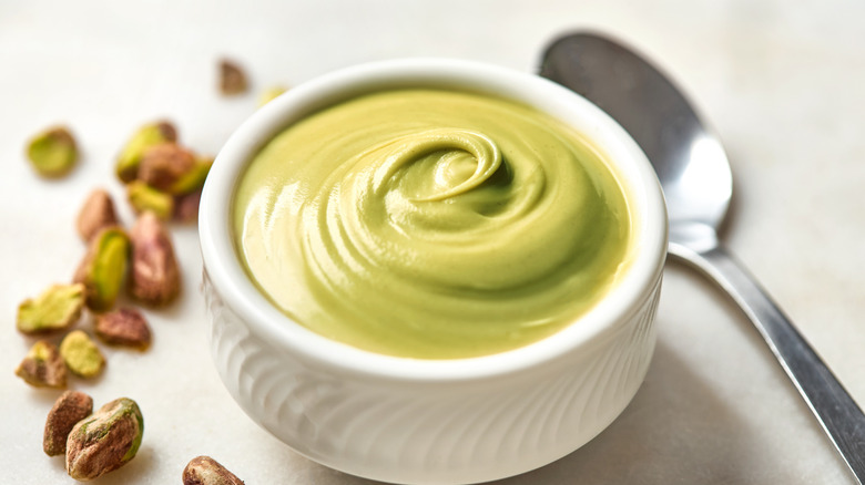 A bowl of pistachio spread with a spoon and pistachio nuts on the table