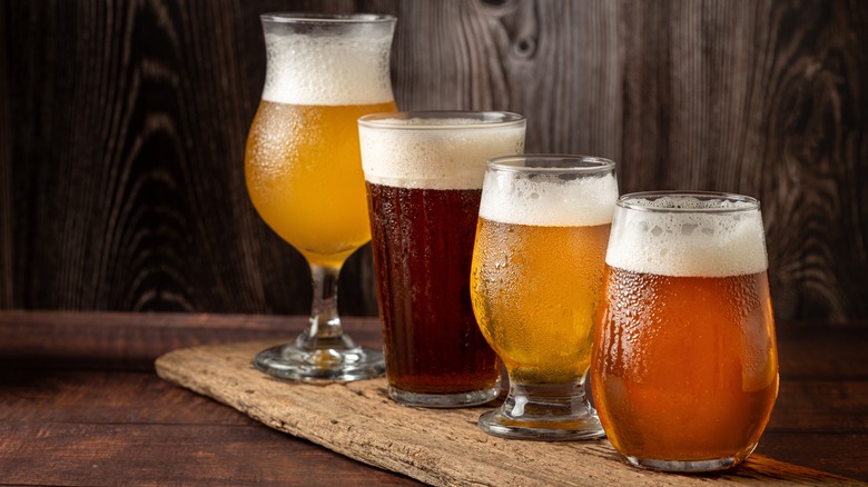 Beers in different golden to brown colors in different shaped glasses on a long light wooden board that's on a dark wood surface