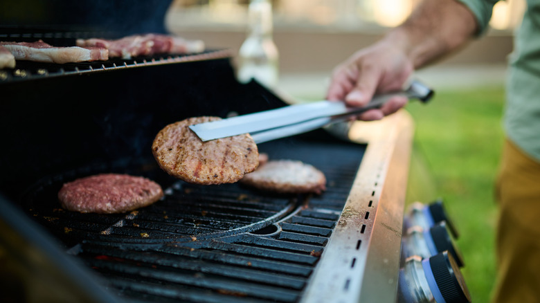 A hand holding tongs flipping burgers on a grill