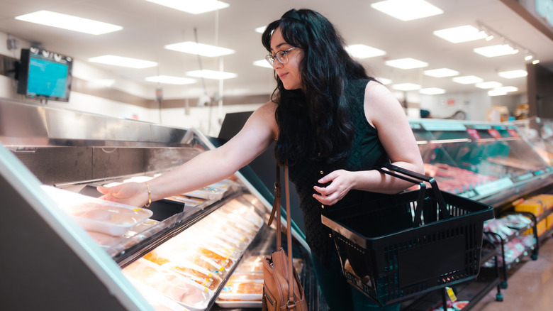 A woman shopping at the grocery store with a hand basket on her arm grabbing a package of chicken meat.