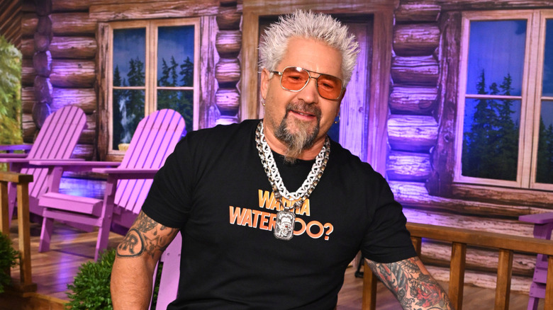 Guy Fieri in front of a fake log cabin in a porch chair