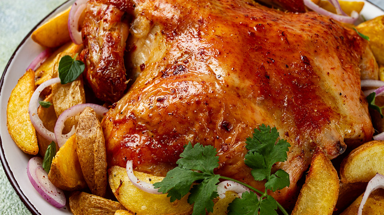 Close-up of golden brown roasted chicken on a round white plate with roasted potato wedges, red onion slices, and a parsley sprig